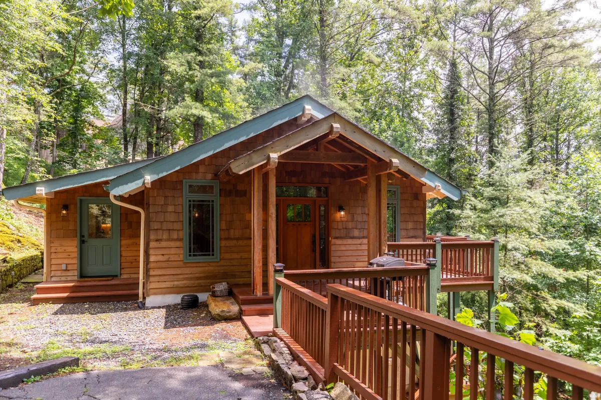 The image shows the welcoming exterior of a single-family mountain home nestled in lush greenery, with a spacious wooden porch at the front. The porch invites relaxation with beautiful natural surroundings, perfect for enjoying sunsets or a cup of coffee in the fresh mountain air. The scene exudes warmth and tranquility, hinting at the peaceful retreat inside.