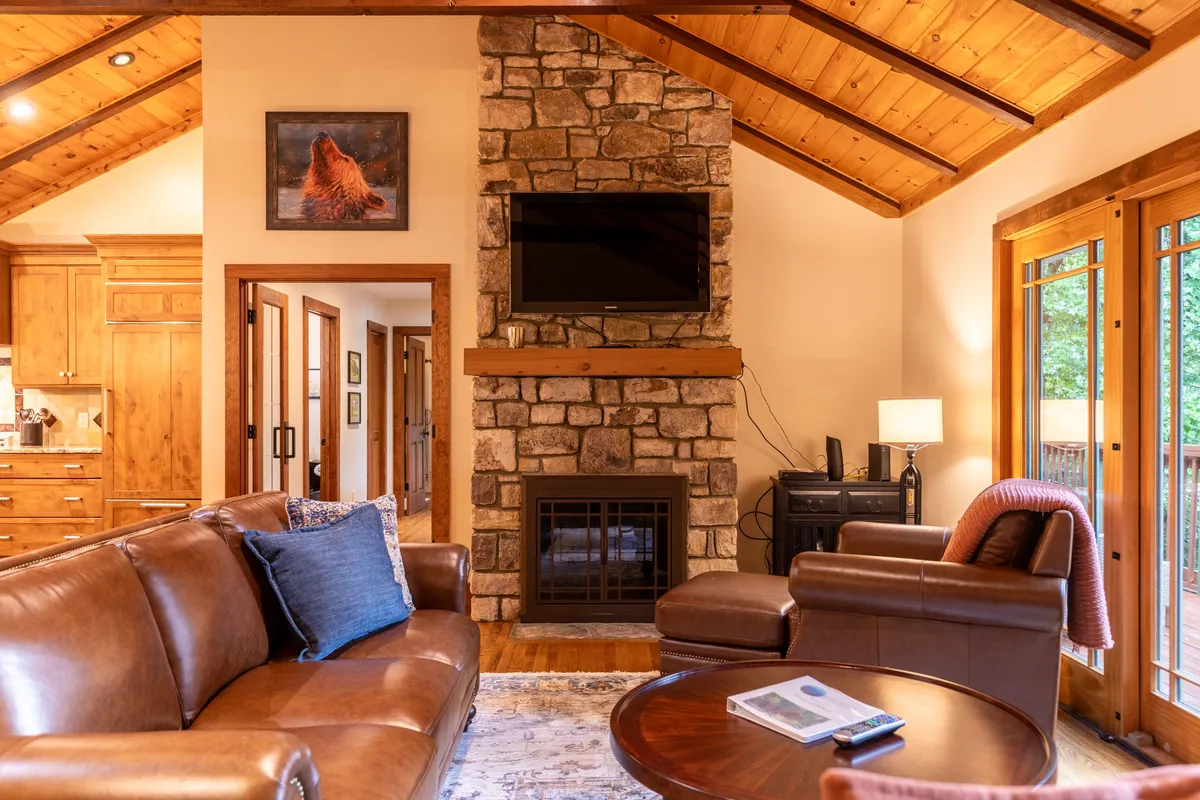 An inviting interior shot with a stone fireplace as its centerpiece, flanked by a leather sofa and wooden beams, with large windows providing natural light and glimpses of the deck beyond.