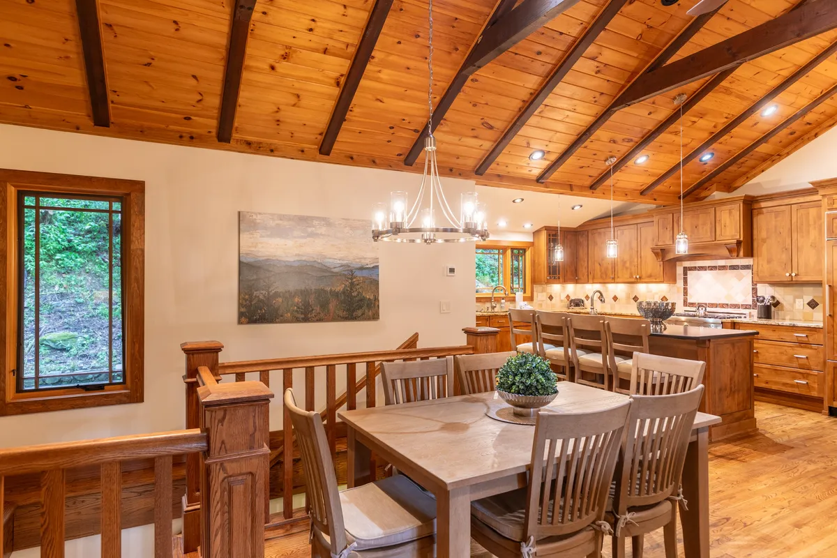 This scene highlights the dining area beneath a chandelier, with wooden beams overhead and a view into the kitchen area. Artwork and soft lighting create a cozy, elegant environment for family meals and gatherings.