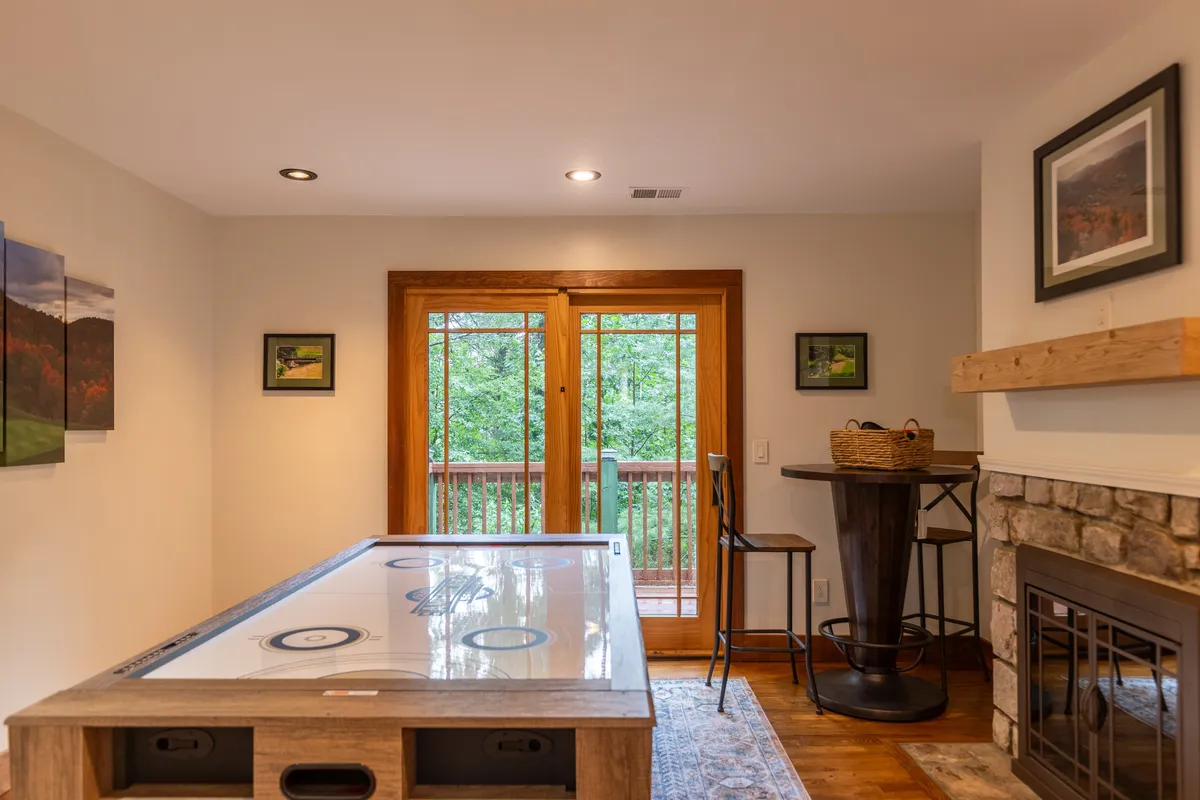 A game and lounge area featuring an air hockey table, a fireplace, and double doors leading out to a sunlit deck. Bar stools invite socializing, and the space seamlessly connects indoor enjoyment with easy outdoor access.