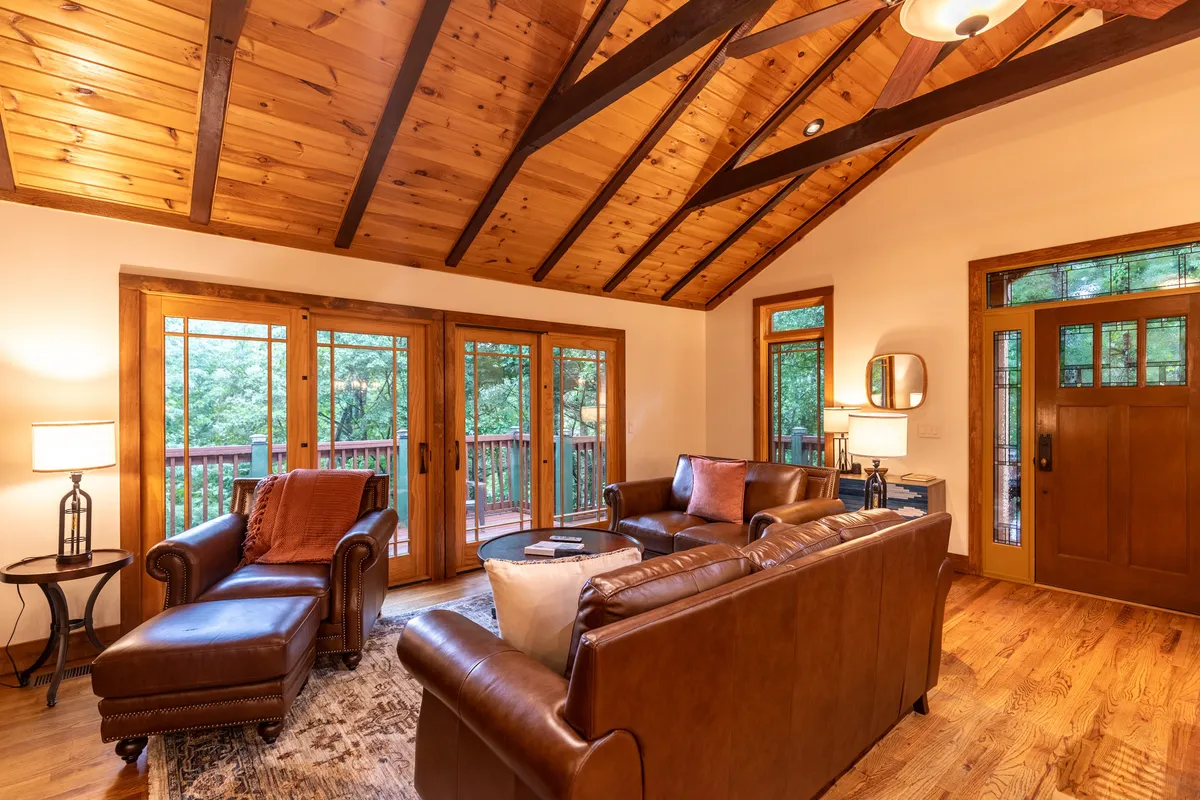 A cozy interior living area with leather sofas arranged for conversation, a warm wooden ceiling, and expansive windows that flood the space with natural light and mountain sunrise views. The two-sided fireplace offers ambiance from multiple vantage points, inviting relaxation.