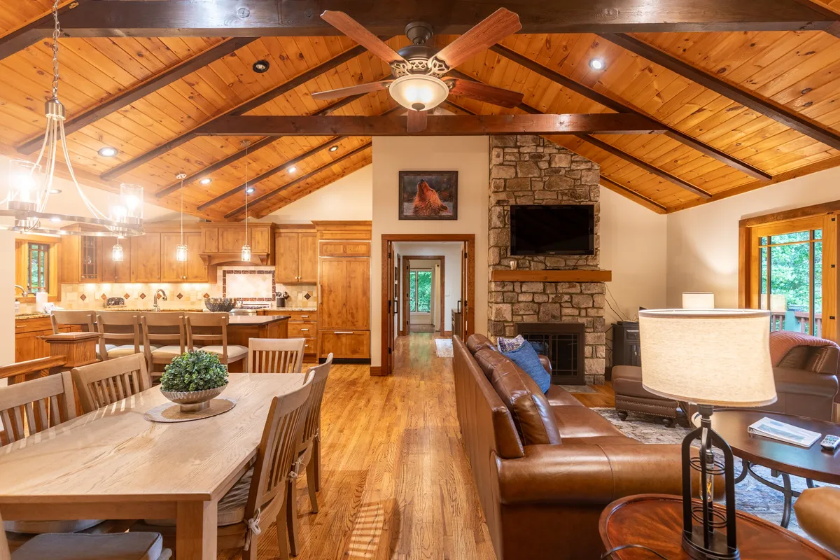 This image presents a welcoming open-concept living and dining area. The focal points include a large wooden dining table, a dramatic stone fireplace, a comfortable leather sofa, and expansive windows that allow natural light to pour in.