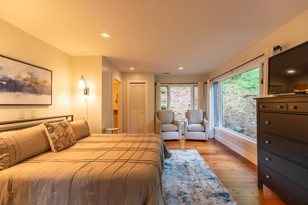 A serene bedroom featuring a plush bed adorned with decorative pillows, flanked by large windows that bathe the space in natural light. Comfortable armchairs and a dresser with a television create an inviting environment for relaxation, reading, or mornings with coffee while enjoying the surrounding views.