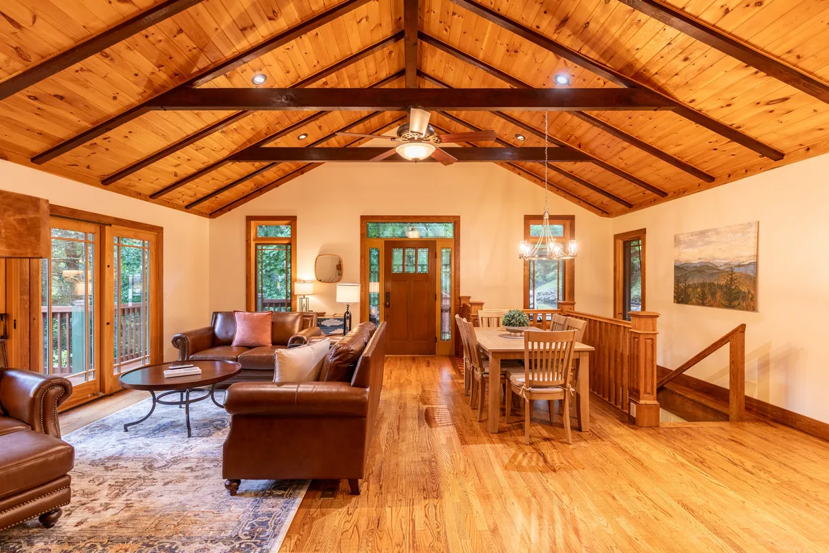 A warmly lit living area featuring exposed wooden beams, a plush sofa set, a dining table ready for gatherings, and expansive windows that offer views of the winter landscape outside. The hearth and the overall ambiance evoke a sense of comfort after chilly outdoor adventures.