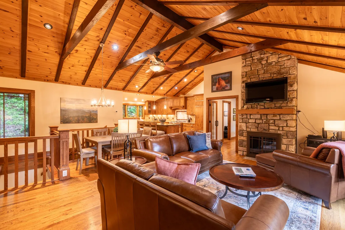 A warm and spacious living room featuring a prominent stone fireplace, plush leather sofas arranged for easy conversation, and exposed wooden beams overhead. A dining area sits in the background, visually linking gathering and relaxation zones.