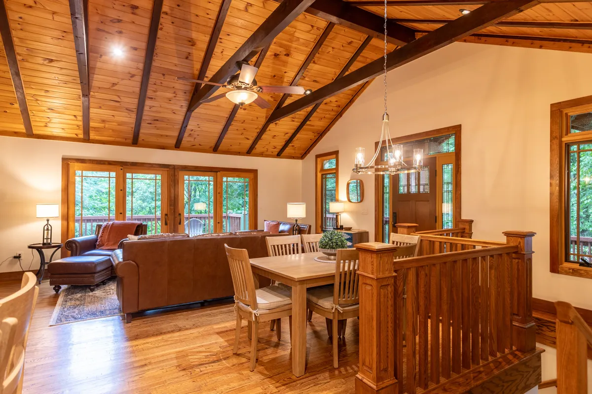 A spacious open-concept living and dining area with wooden ceiling beams, a large leather sofa, a dining table set for festive meals, and generous windows revealing glimpses of natural surroundings—suggesting a seamless connection to the outdoors.