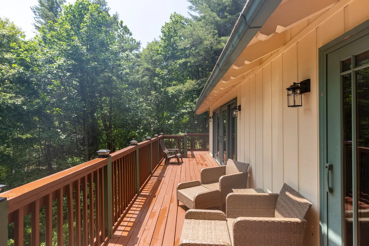 This image features an inviting outdoor deck surrounded by tall trees, with comfortable seating arranged to overlook the woodland view. The warm light suggests either morning or late afternoon, and a tranquil atmosphere prevails, perfect for relaxing, enjoying nature, or reading.